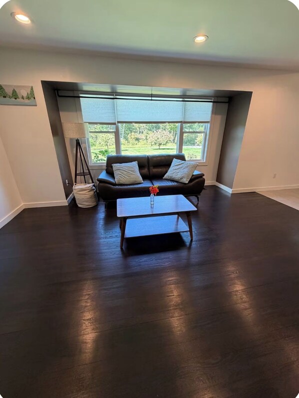A room with a dark wood floor, a sofa with cushions, a coffee table, and a window with blinds.