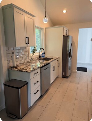 A modern kitchen with white cabinets, a granite countertop, a stainless steel refrigerator, and a compact trash bin.