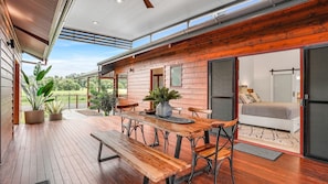 A wooden deck with a dining table and chairs, a potted plant, and a bedroom with a bed and a sliding door.