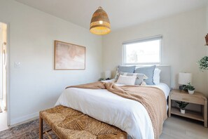 Ein Schlafzimmer mit Bett, Nachttischen, einer Pendelleuchte, einem Fenster und einem an der Wand befestigten Kunstwerk.