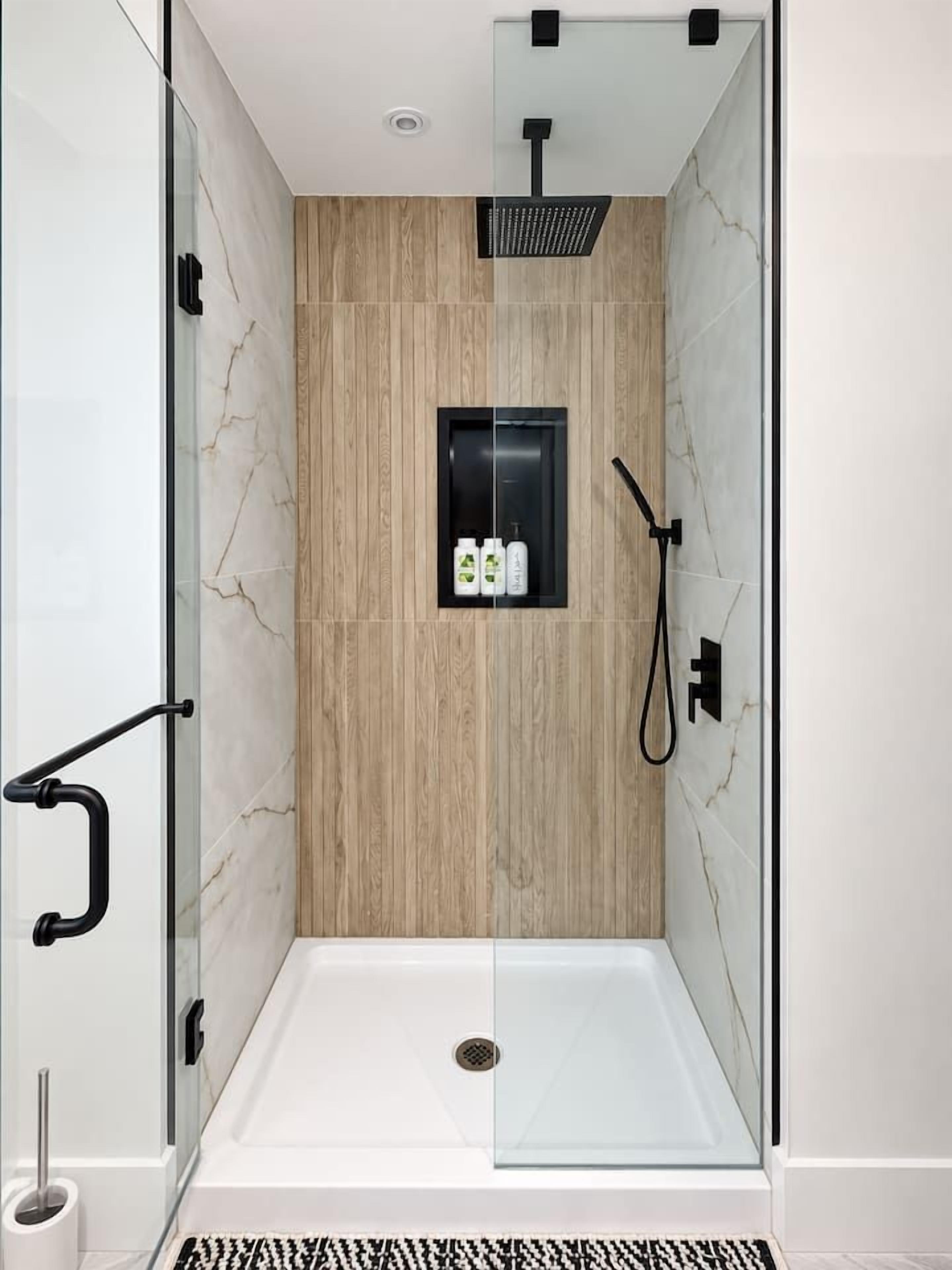 A glass-enclosed shower stall with a wooden panel wall, a black handheld showerhead, and a small shelf with toiletries.