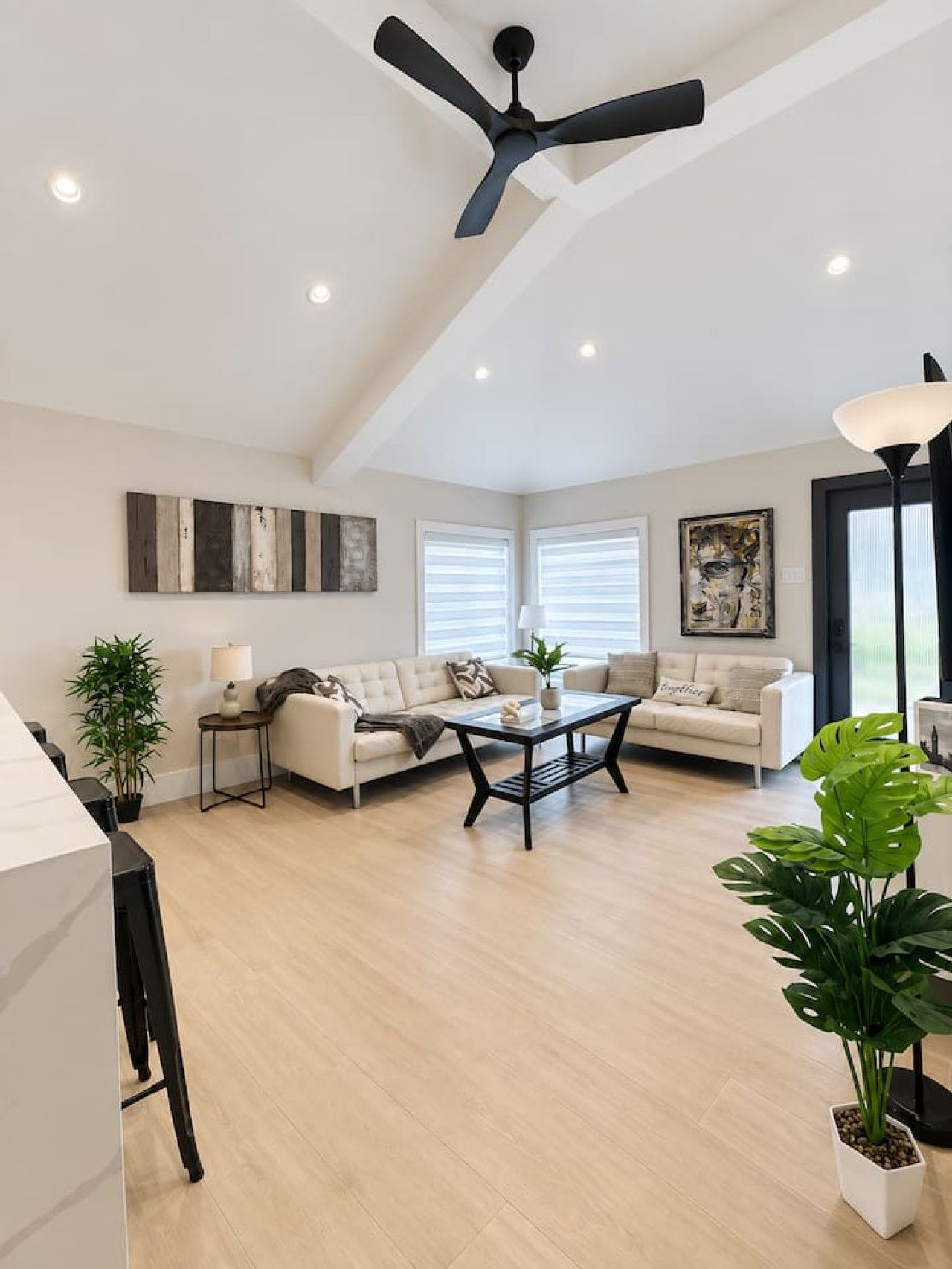 A spacious living room with a high ceiling, a ceiling fan, and a large window with blinds. There are two sofas, a coffee table, and a potted plant.