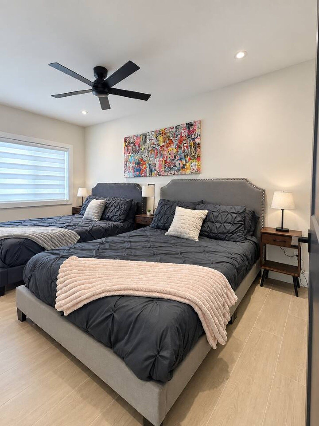 A bedroom with two beds, a ceiling fan, a colorful artwork on the wall, and a bedside table with a lamp.
