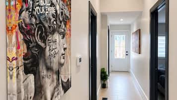 Un pasillo con un cuadro abstracto de gran tamaño en la pared, una puerta al final y una planta en maceta en el suelo.