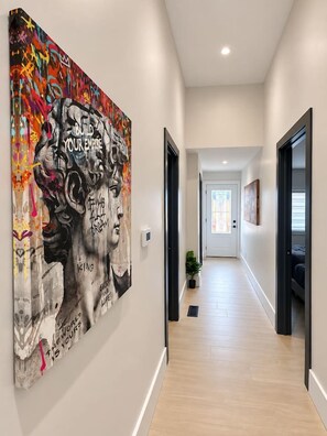 Un couloir avec une grande peinture abstraite au mur, une porte au fond et une plante en pot posée sur le sol.