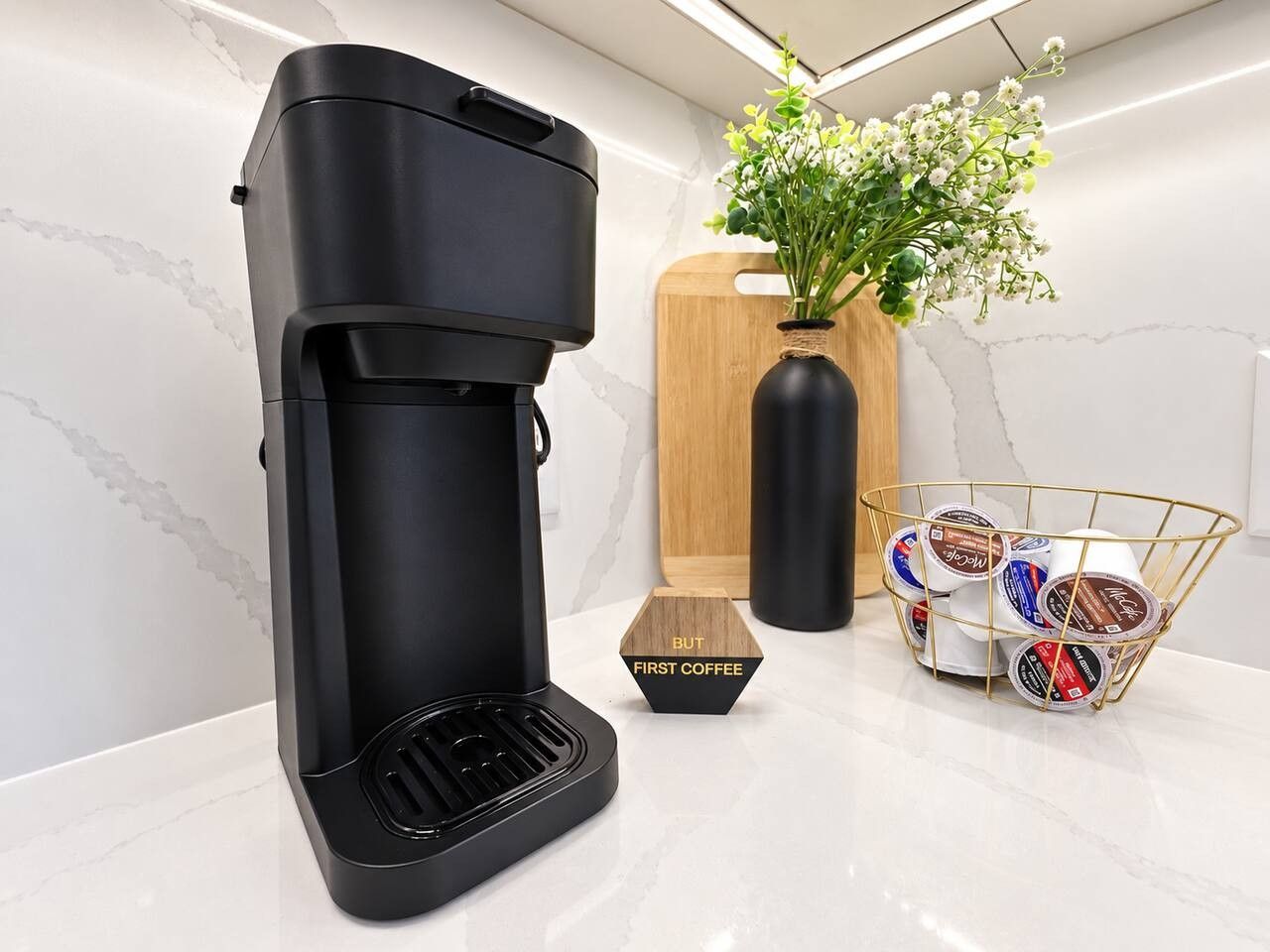 A black coffee maker on a white countertop with a wooden cutting board, a vase with flowers, and a basket of coffee pods.