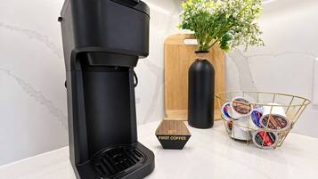 A black coffee maker on a white countertop with a wooden cutting board, a vase with flowers, and a basket of coffee pods.