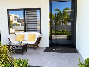 A modern outdoor seating area with a sofa and a glass-top table, a sliding glass door, and a doormat with the name 'BLISSFUL ABODE'. - Blissful Abode/ Beach/Ps5/Courtyard Oasis/ Pool (Llandovery)