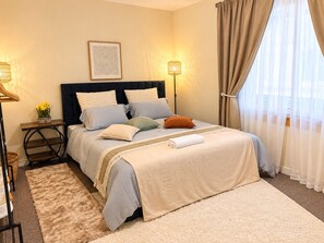 Un dormitorio ordenado con cama, mesitas de noche, alfombra y ventana con cortinas.