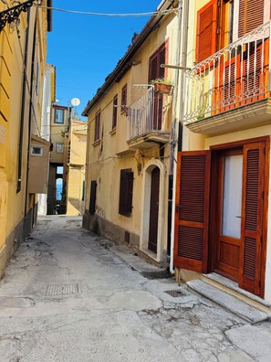 A narrow street with buildings on both sides, featuring balconies with railings and wooden shutters.