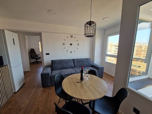 Un espace de vie moderne avec une table ronde en bois, des chaises noires, un canapé gris et une grande fenêtre donnant sur des immeubles.
