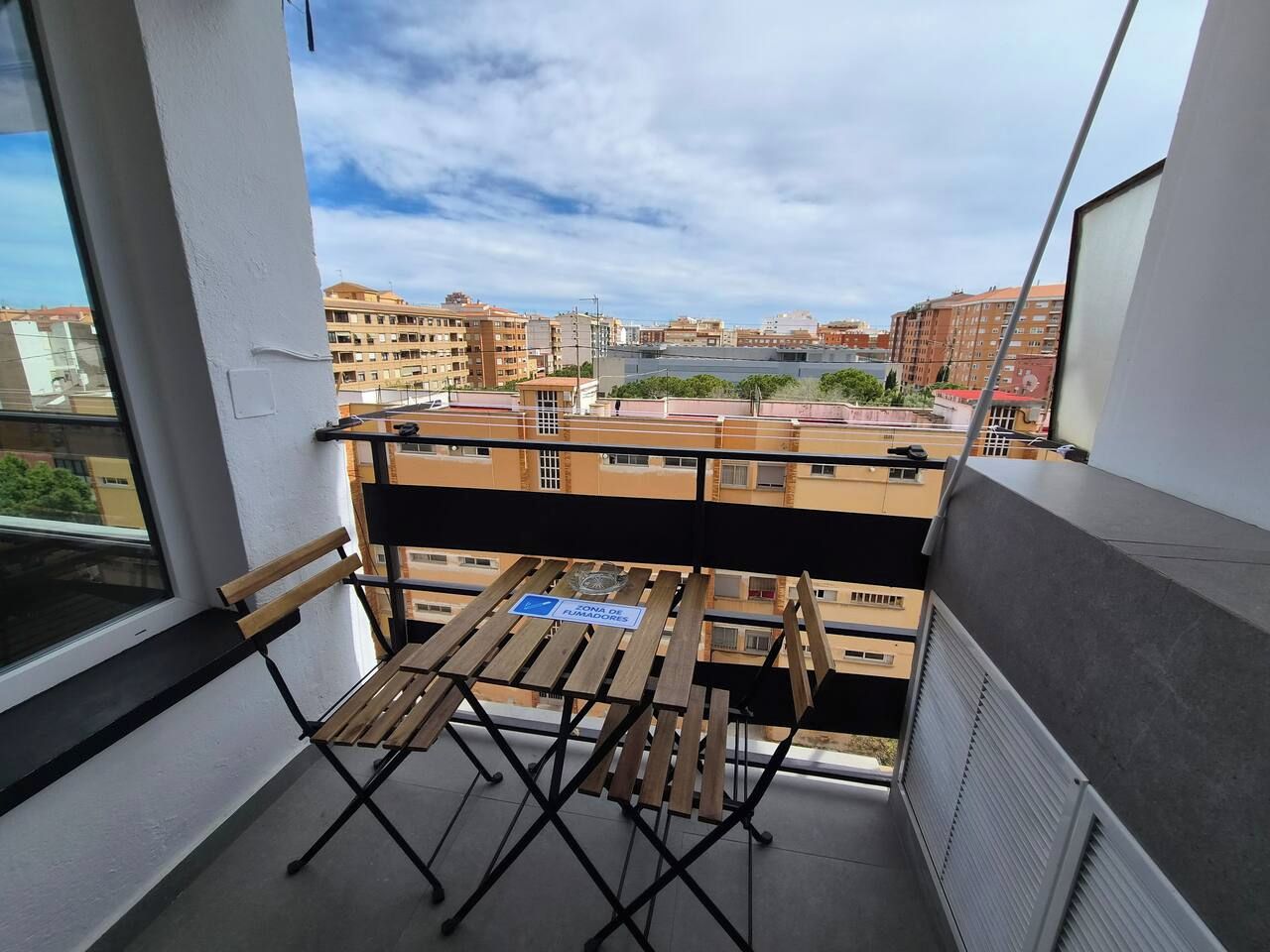 Ein Balkon mit Holztisch und Stühlen, ein Schild mit der Kennzeichnung Raucherbereich sowie Blick auf die umliegenden Gebäude.