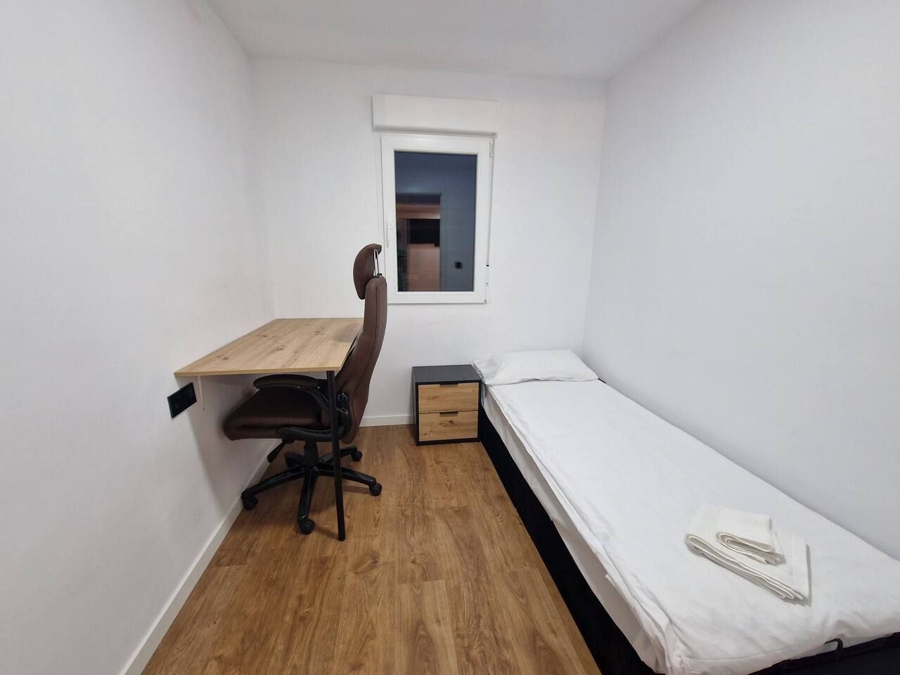 Ein kleines Zimmer mit einem Holztisch, einem Stuhl, einem Einzelbett und einem kleinen Fenster.
