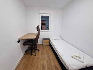 Quarto pequeno com escrivaninha de madeira, uma cadeira, uma cama de solteiro e uma janela.