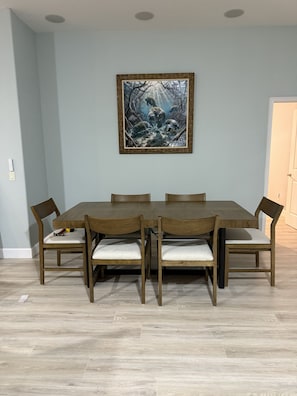 Ein Essbereich mit einem rechteckigen Holztisch und sechs Stühlen, einem gerahmten Bild an der Wand und einem hellen Boden.