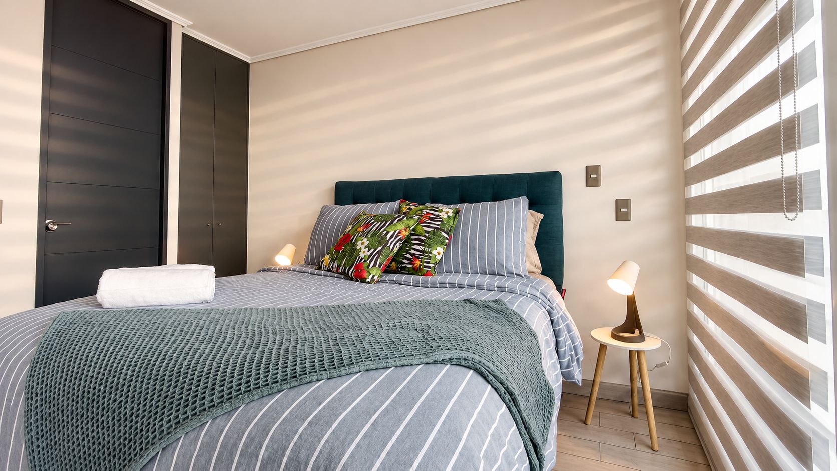 A neatly made bed with a striped bedspread and pillows, a bedside lamp, and a view of a striped wall.