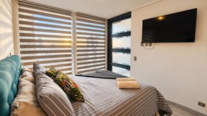 A hotel room with a bed, a sofa, a TV mounted on the wall, and a window with blinds.