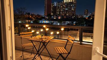 A balcony with a wooden table and chairs, string lights, and a view of a cityscape at night.
