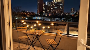 A balcony with a wooden table and chairs, string lights, and a view of a cityscape at night.