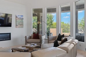 En moderne stue med sofa, sofabord og fjernsyn. Store vinduer giver udsigt over grønne områder, og der er en balkon.