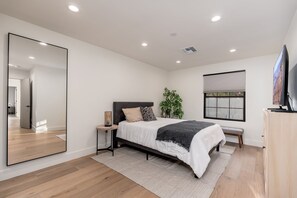 Een moderne slaapkamer met een bed, nachtkastjes, een spiegel, een raam met zonwering en een televisie.