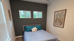 Quarto com cama, ventilador de teto, duas janelas e uma obra de arte emoldurada na parede.