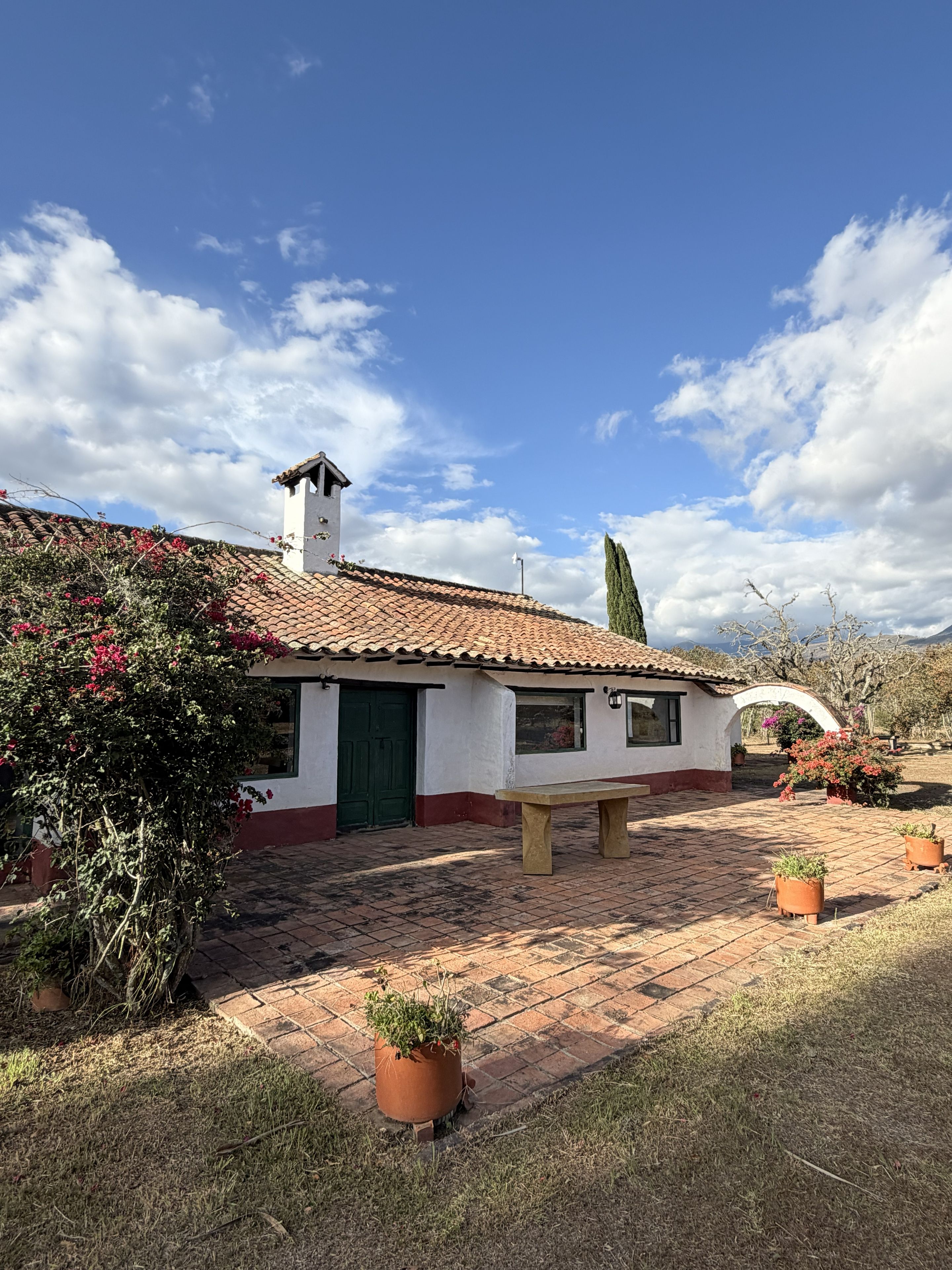 A white building with a tiled roof, a green door, and a bench in front. There are potted plants and a clear sky with clouds.