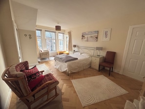 Un dormitorio con cama, una silla, una mesita de noche, una lámpara y un ventanal.