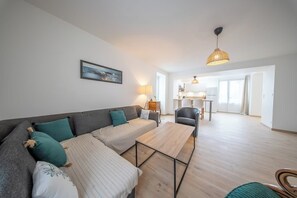 A modern living room with a sofa, a coffee table, and a dining area in the background. - Petit coin de paradis - charmante maison jardin (Quiberon)
