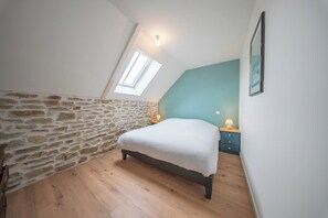 Une chambre avec un mur en pierre, un plafond en pente, un lit, une table de chevet et une fenêtre.