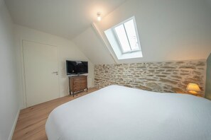 A bedroom with a sloped ceiling, a skylight, a bed, a dresser, a television, and a stone wall. - Petit coin de paradis - charmante maison jardin (Quiberon)