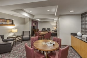 Une chambre d’hôtel avec une table de salle à manger ronde, des chaises à revêtement rouge, une petite table d’appoint avec une lampe, un coin cuisine et un tapis à motifs.