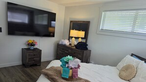 A hotel room with a bed, a dresser, a TV, a window with blinds, and a nightstand with a vase of flowers. - My Paradise, a 4-bedroom Home with Hot Tub & Swimming Pool in Soothing Pensacola (Pensacola)