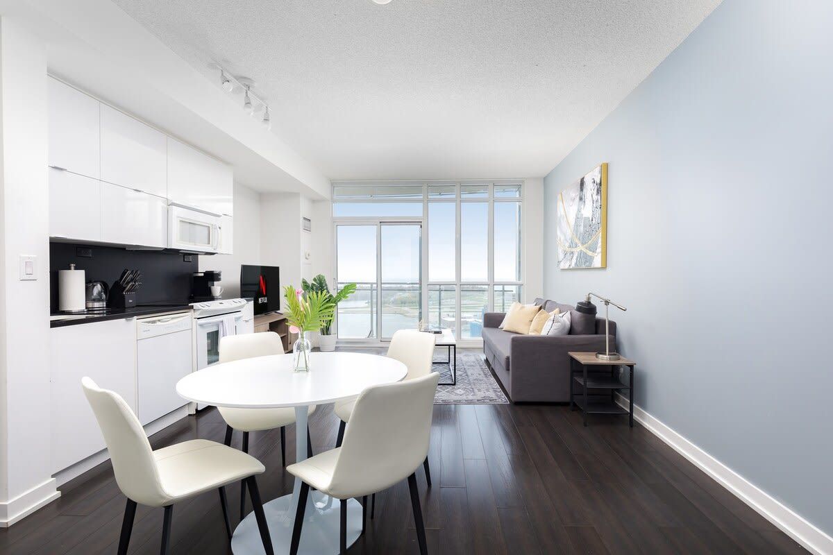 A modern kitchen with white cabinets, a dining area with a white table and chairs, and a gray sofa in a living room with a large window.