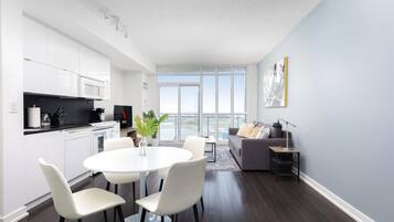 A modern kitchen with white cabinets, a dining area with a white table and chairs, and a gray sofa in a living room with a large window.