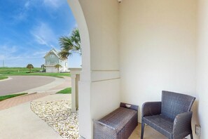A balcony with a wicker chair and a bench, overlooking a road and a house. - Palmilla Beach Resort-137 Palmilla Circle Unit 104 (Port Aransas)