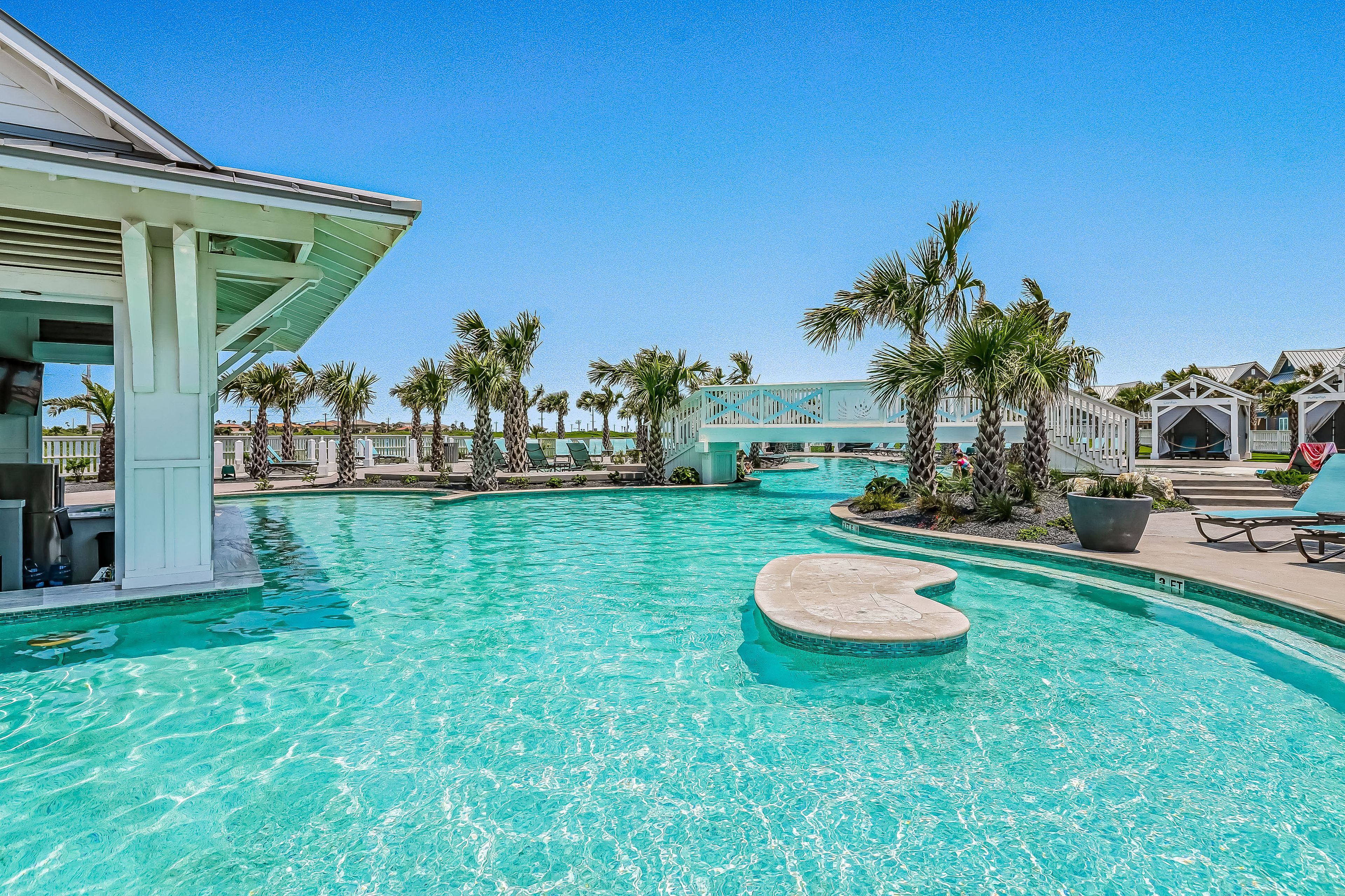A clear blue pool with a central island, surrounded by palm trees and lounge chairs.