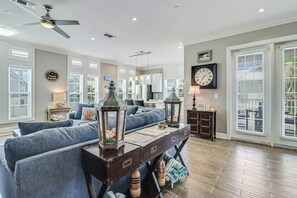 A spacious living room with a ceiling fan, a clock, and a view of the outdoors through large windows. - Palmilla Beach Resort-137 Palmilla Circle Unit 201 (Port Aransas)