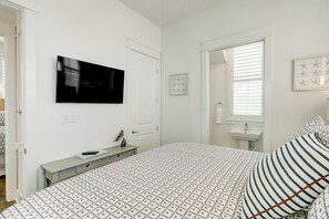 Ein Schlafzimmer mit einem Bett, einem an der Wand montierten Fernseher, einem Nachttisch, einem Fenster mit Jalousien und einem kleines Badezimmer mit einem Waschbecken.