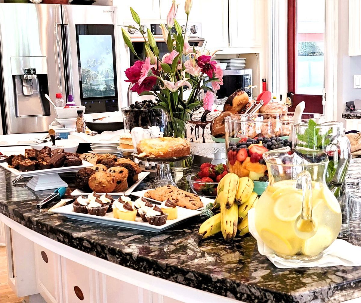 A kitchen counter with a variety of food items, including pastries, fruits, and a pitcher of lemonade.