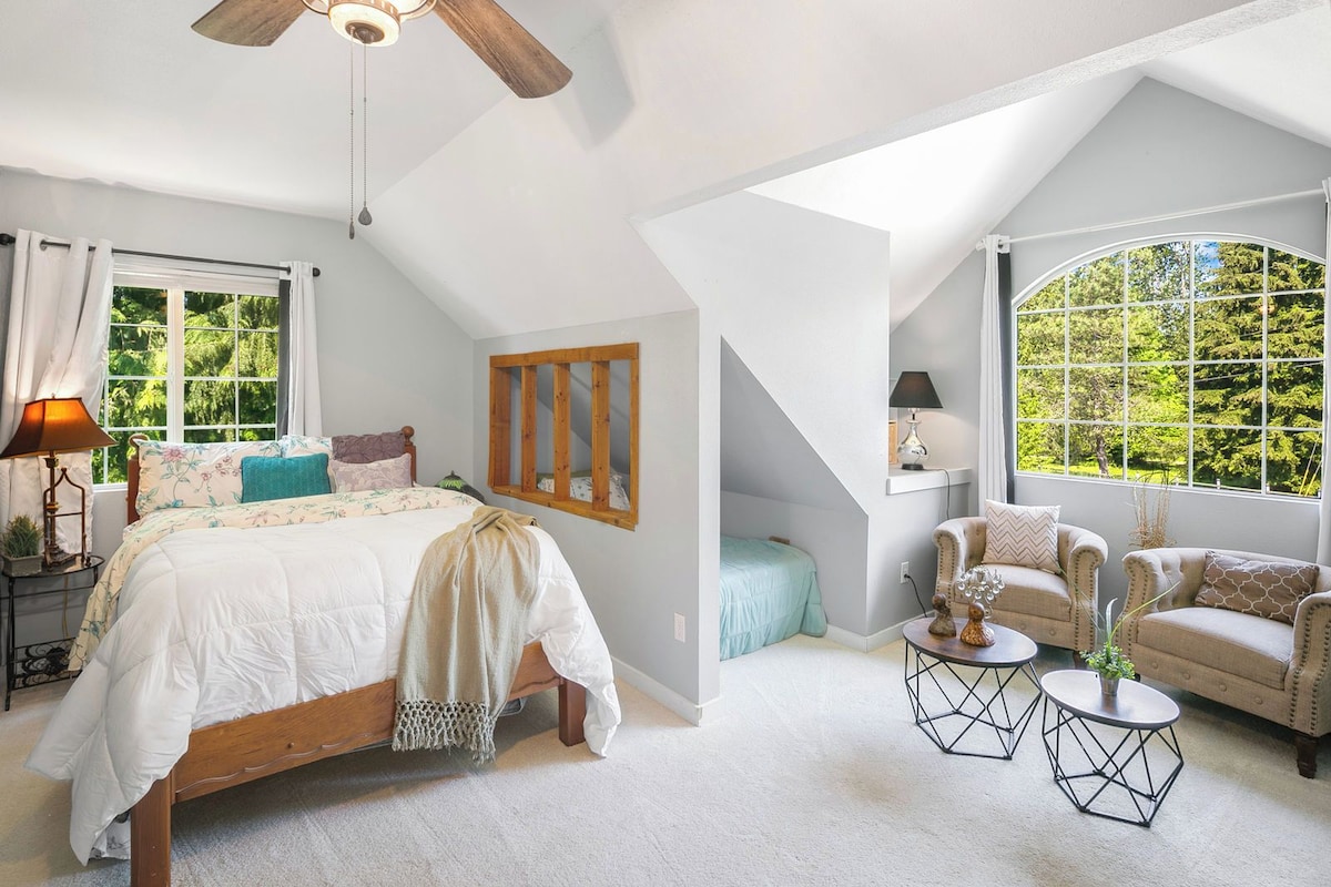 A bedroom with a bed, two chairs, a small table, a window, and a ceiling fan.
