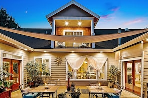 A two-story house with a covered patio area featuring multiple tables and chairs, surrounded by potted plants.