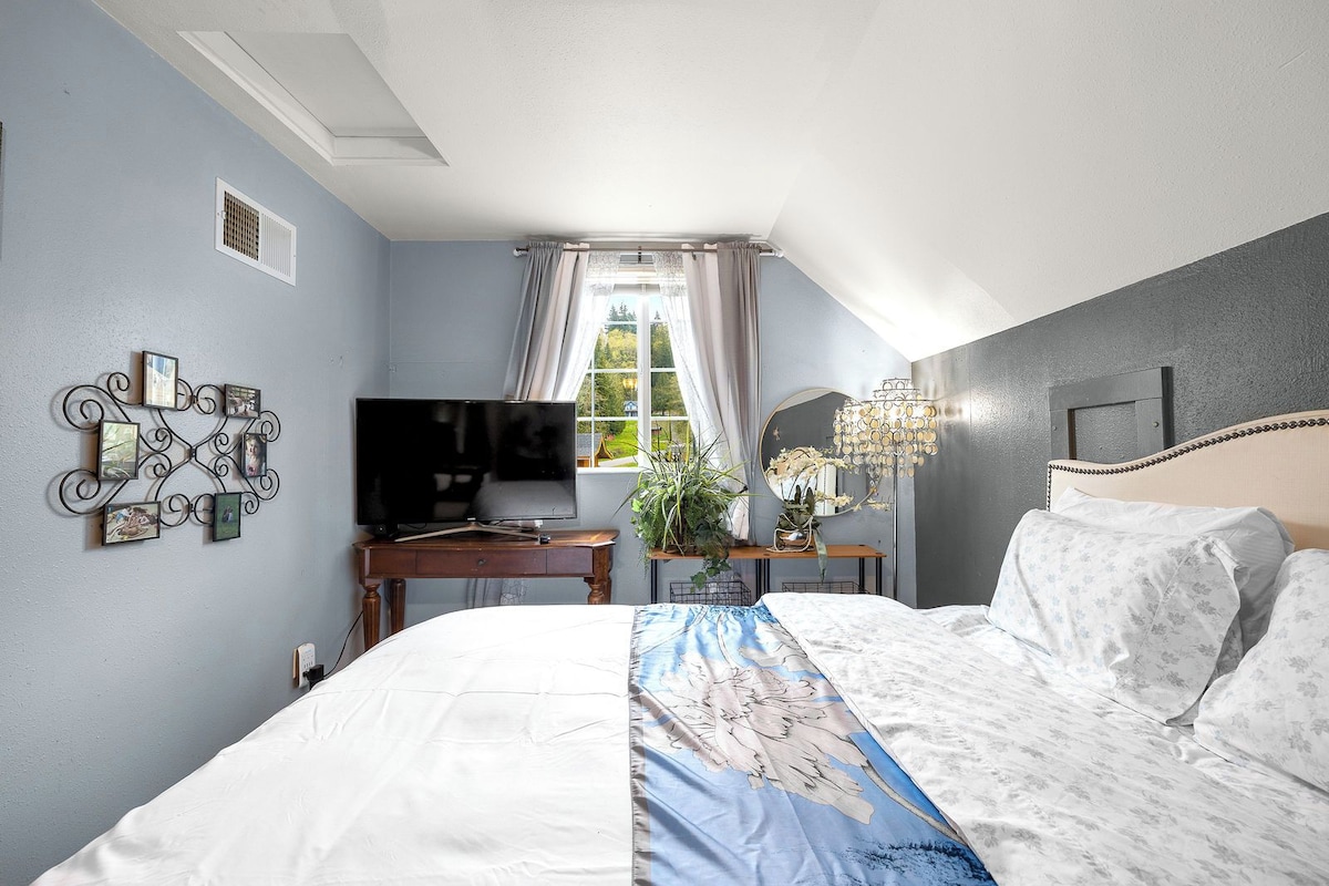 A bedroom with a bed, a television on a desk, a window with curtains, and a decorative wall piece.