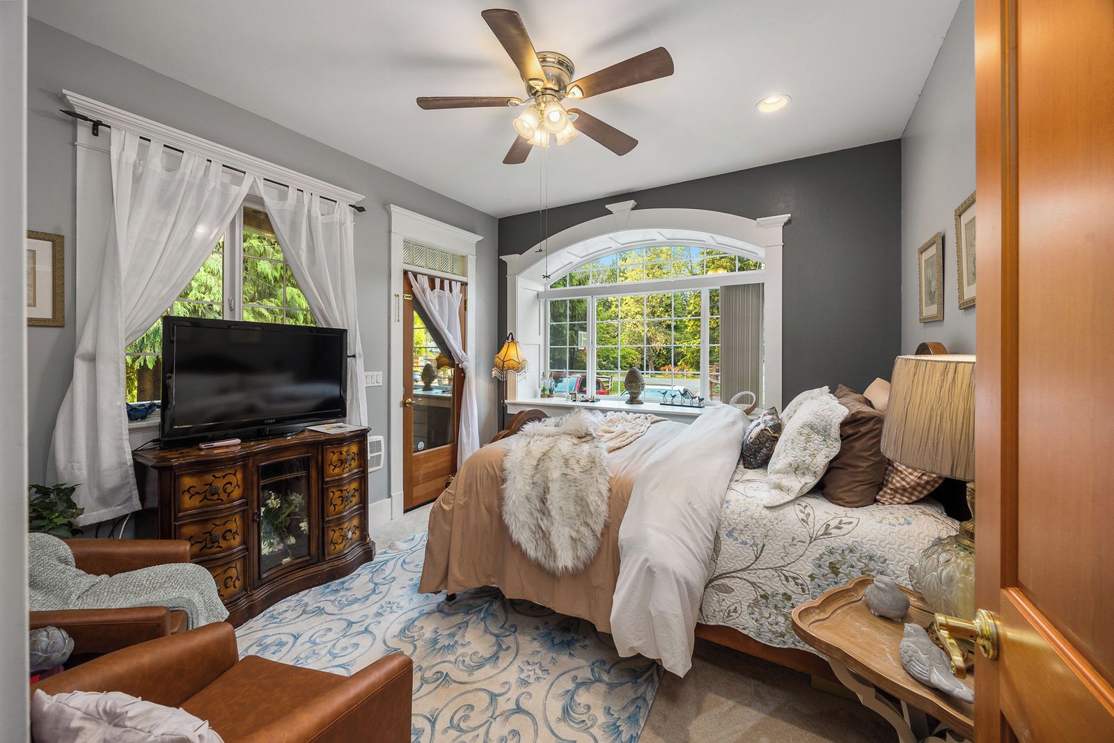 A bedroom with a bed, a television on a wooden cabinet, a ceiling fan, and a window with a view of greenery.