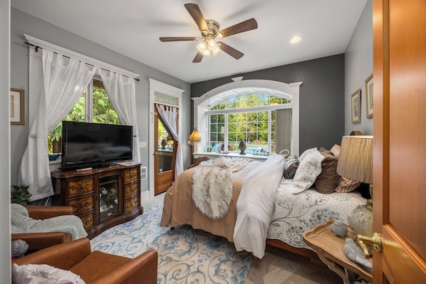 A bedroom with a bed, a television on a wooden cabinet, a ceiling fan, and a window with a view of greenery.