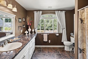 A bathroom with a marble countertop, a white sink, a toilet, and a window with curtains. - The Grayson B&B Extended Stays (Monroe)