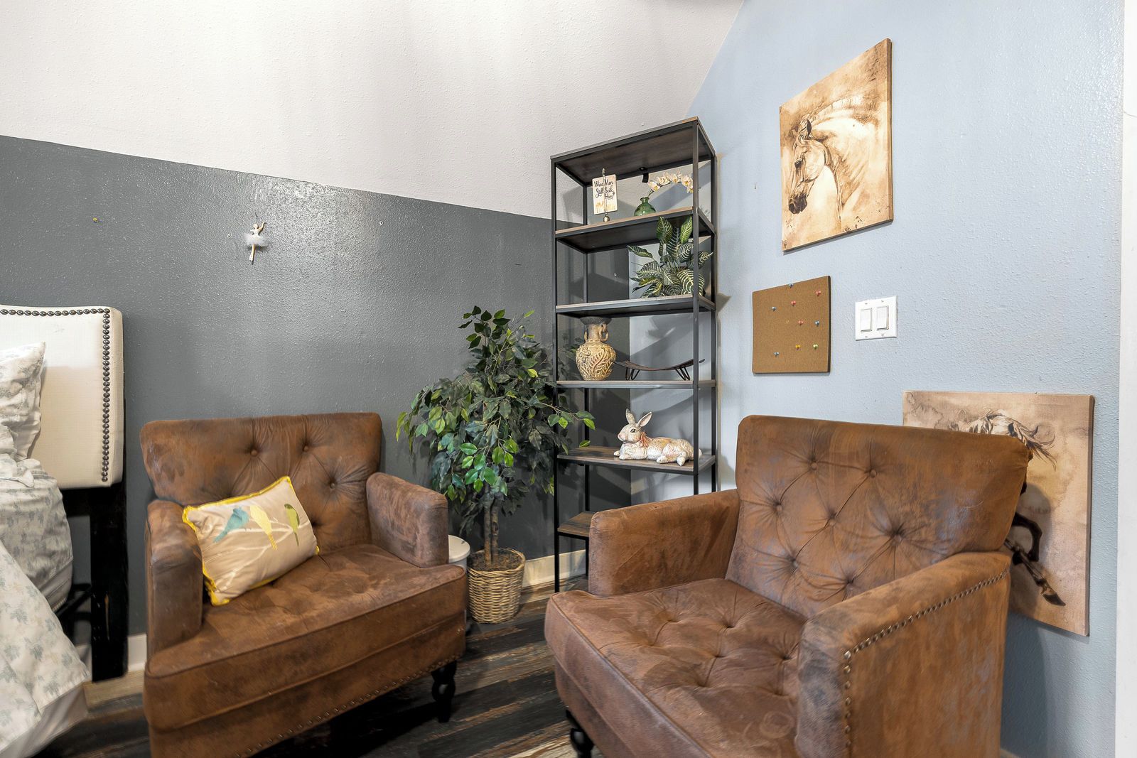 A room with two brown leather armchairs, a black metal shelving unit with decorative items, a potted plant, and a wall-mounted artwork.