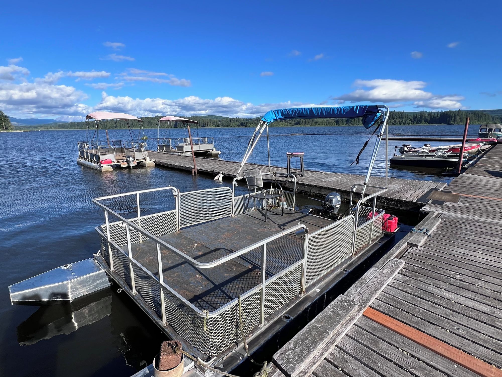 Un muelle con un barco, un área de estar cubierta y vistas a un lago con montañas al fondo.