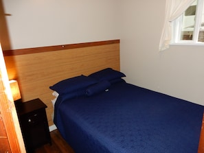 A single bed with a wooden headboard and a blue bedspread in a room with a window and a nightstand.