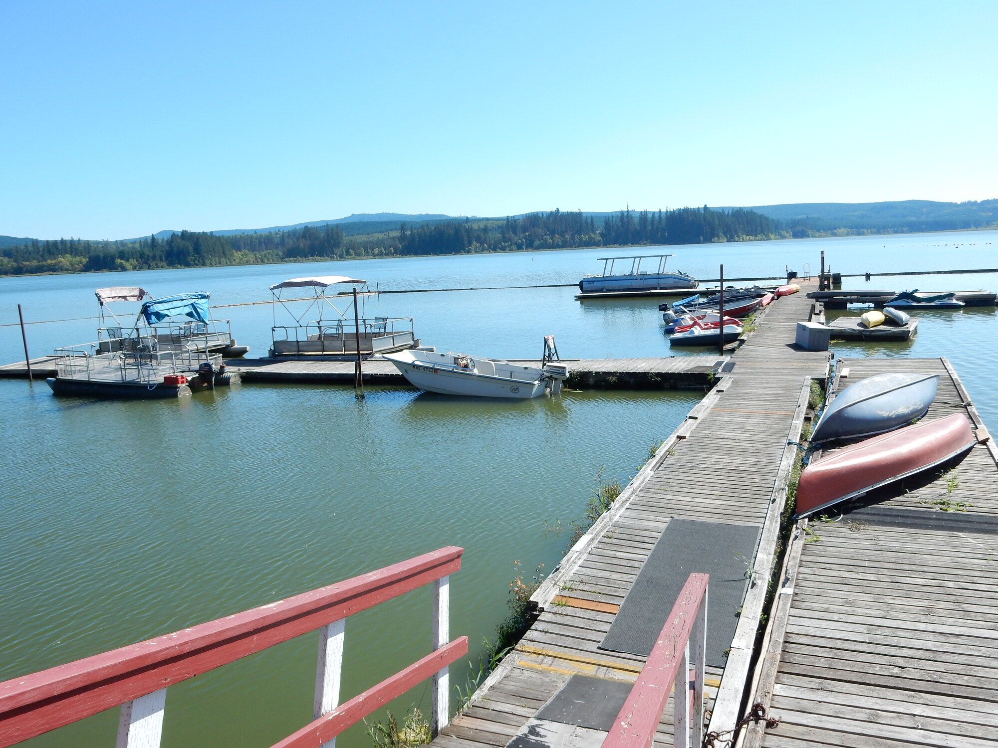 A wooden dock with boats moored at the end, a clear sky, and a calm lake with a forested shoreline in the background.